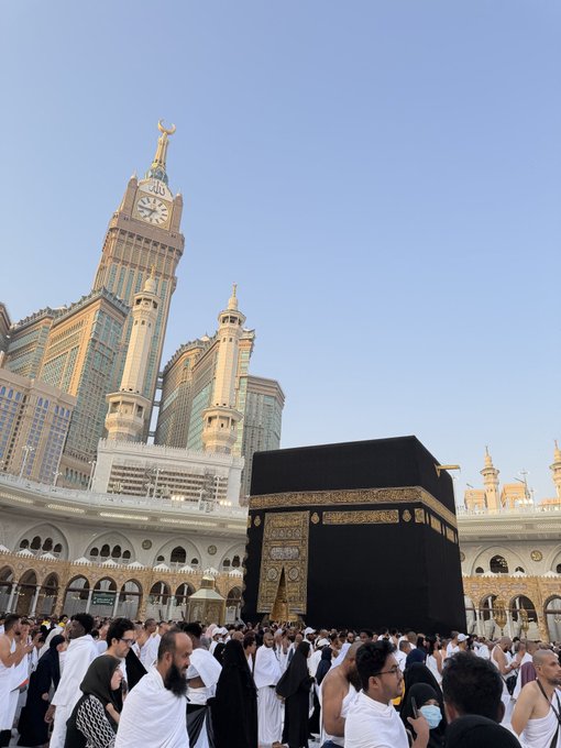 A crowd of pilgrims gathers around the Kaaba at the Masjid al-Haram in Mecca, with the iconic Abraj Al-Bait clock tower and golden crescent visible in the background.