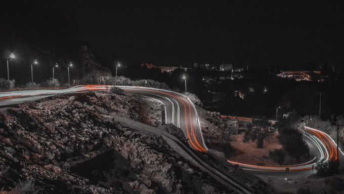 A nighttime long-exposure photograph showing light trails from vehicles winding along a curved mountain road illuminated by streetlights.