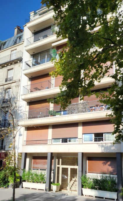 A multi-story residential apartment building with balconies and reddish-brown window shutters is partially obscured by green tree foliage in the foreground.