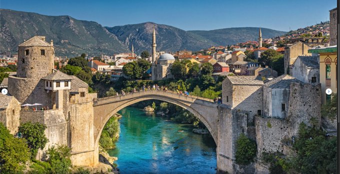 A scenic view of the Stari Most bridge in Mostar, Bosnia, spanning a turquoise river with historic buildings and mountains in the background.