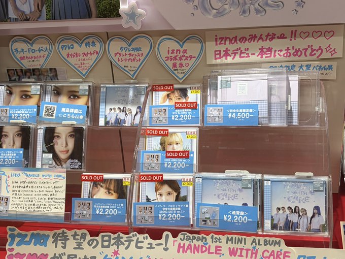 A Japanese retail display showcases IZ*ONE's debut mini album "HANDLE WITH CARE," with multiple versions marked "SOLD OUT" and priced at ¥2,200 (¥4,500 for the full set), alongside handwritten fan signs supporting the group's Japan debut. The note "COCOXIM also SOLD OUT" appears handwritten on a heart-shaped card on the upper shelf, indicating that even the limited COCOKIM variant is no longer available, reflecting the album's overwhelming popularity.