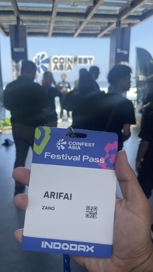 A hand holds a 'COINFEST ASIA Festival Pass' with the name 'ARIFAI' and 'ZANO', with 'INDODAX' printed at the bottom, set against a blurred background of event attendees.