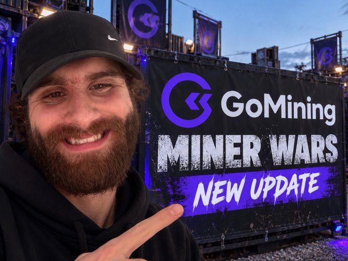 A smiling bearded man in a black Nike cap and hoodie points at a large GoMining sign reading “MINER WARS” and “NEW UPDATE.”