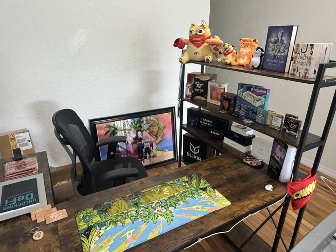 A wooden desk setup featuring a black office chair, a colorful illustrated desk mat, framed artwork leaning against the wall, and a shelf displaying various figurines, merchandise, and a small ELGATO box.