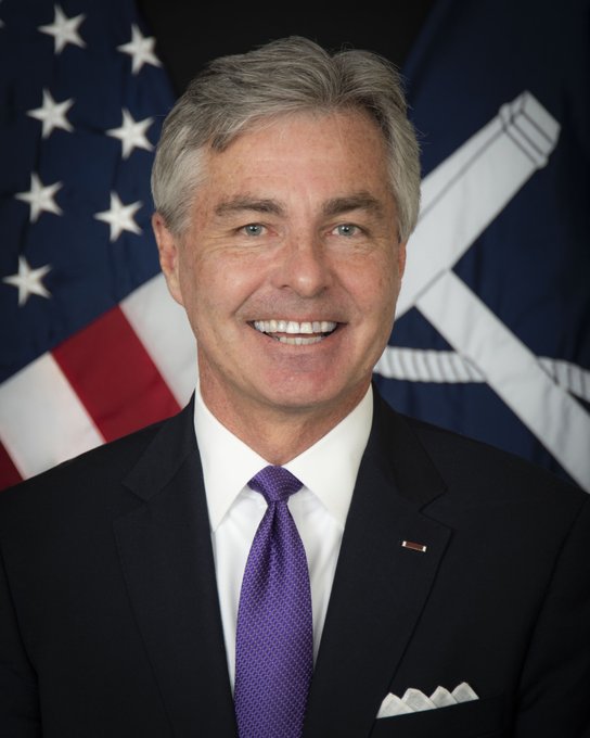 An official portrait of a smiling man with gray hair wearing a dark suit, white shirt, and purple tie, posed in front of an American flag and a dark flag with an emblem, with a small American flag pin on his lapel.