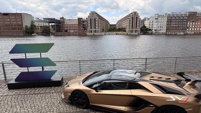 The image features a tan sports car parked next to a body of water with buildings in the background. The car appears to be a Lamborghini Huracan, which has a unique design that sets it apart from other vehicles. The photo also includes text that reads "Solana summit Germany today here in Berlin. Cool location. This is not my lambo btw, i know what i hold."