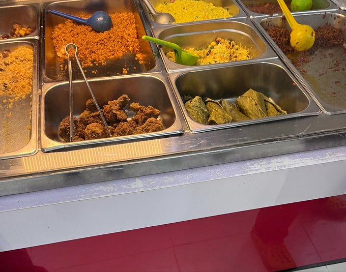 A cafeteria-style buffet serving station with metal trays containing various foods including stuffed grape leaves, rice dishes, and meat, with serving utensils.