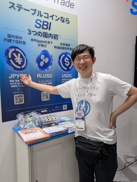 A man wearing a "ステーブルコイン 1対1 ドル" t-shirt poses at the SBI trade booth, which advertises "国内初の円建て預託型ステーブルコイン SBI 3つの国内初" featuring logos for JPYS, RULSD, and USDC, alongside a display of promotional candy being handed out.