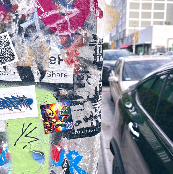 Street photography showing a sticker-covered pole in the foreground featuring a Pepe the Frog sticker and various graffiti tags, with parked cars and a building blurred in the background.