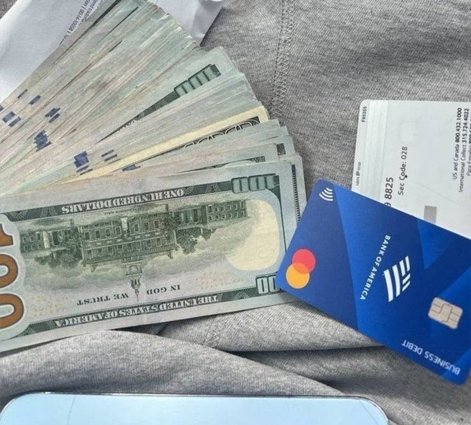 A fan of one-hundred-dollar bills sits on a fabric surface next to a Bank of America Business Debit Mastercard and an Air Canada boarding pass, illustrating the cycle of earning and spending money tied to work and travel.