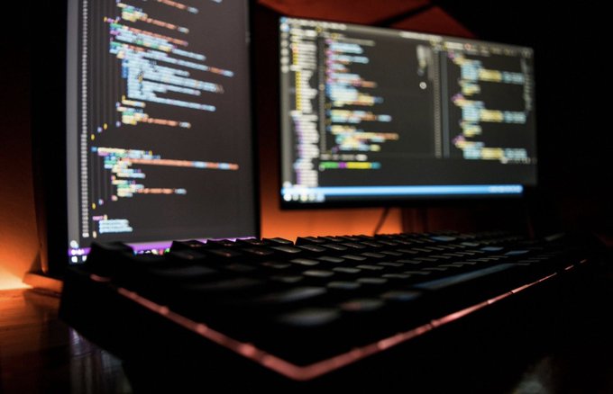 A low-angle close-up of a backlit keyboard in front of two computer monitors displaying lines of colorful code.