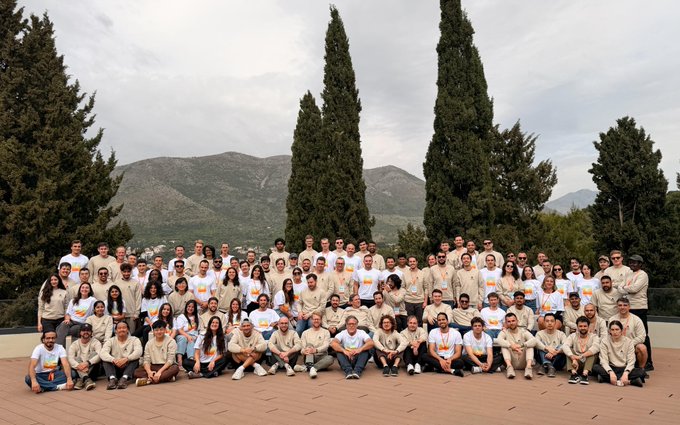 A large group photo of dozens of people wearing matching beige and white shirts, posed outdoors with tall cypress trees and mountains in the background.