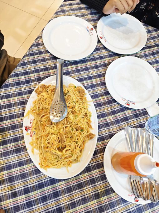 The image shows a table with plates of food, including spaghetti and meatballs, as well as utensils like forks and spoons. The table also has bottles of sauce nearby. There are two people present at the table, one on each side, possibly enjoying their meal together.