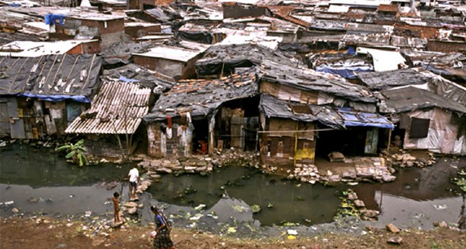 An aerial view shows a slum with dilapidated structures and tarps lining a murky waterway, with a few people standing on the muddy banks.