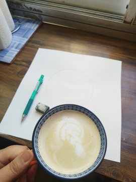 An overhead view of a wooden table by a window with a hand holding a blue-and-white ceramic cup of latte art, alongside a pen, eraser, and a blank piece of paper with a faint pencil circle.