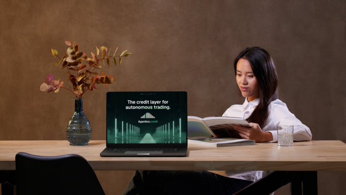 A woman sits at a wooden desk reading a book, with a laptop displaying 'The credit layer for autonomous trading' and the Agentica Credit logo, alongside a vase with autumn foliage.
