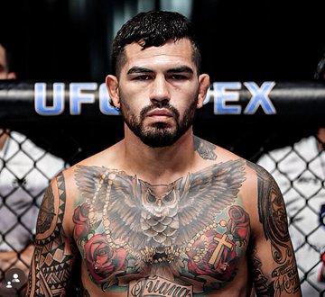 A shirtless UFC fighter with extensive chest and arm tattoos standing in front of a chain-link fence with the 'UFC' and 'APEX' logos visible.