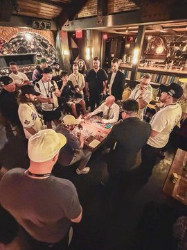 A crowded gathering in a dimly lit venue with brick walls, where people are gathered around a table with a birthday cake, with one person wearing a cap with text that includes 'PATEK' visible.