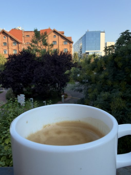 A close-up of a white ceramic mug filled with coffee, set against a background view of buildings, trees, and greenery from what appears to be a balcony or window.