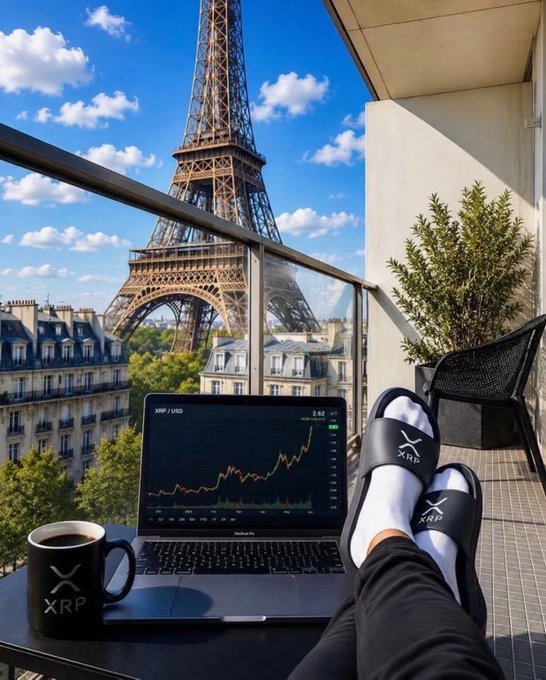 A first-person view of feet in XRP-branded slides resting near a laptop displaying an XRP/USD chart, with the Eiffel Tower visible in the background alongside an XRP mug.