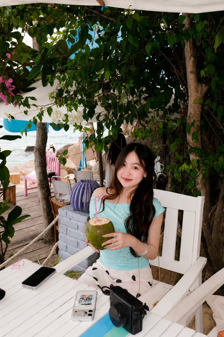 A young woman with long dark hair sits at an outdoor table holding a fresh coconut with a straw, with the ocean visible in the background.