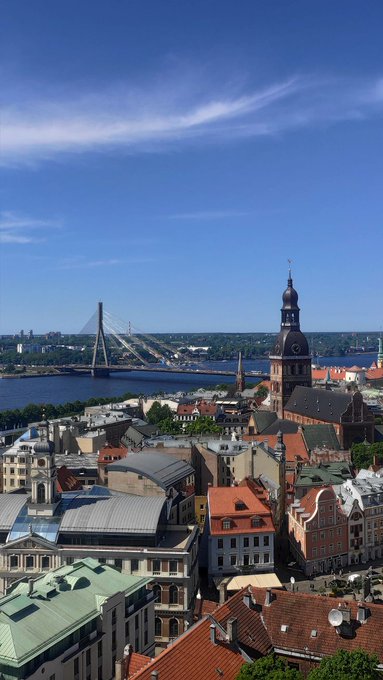 A cityscape featuring a river spanned by a distinctive cable-stayed bridge, with a prominent church spire and a mix of older buildings with red roofs in the foreground under a bright blue sky.
