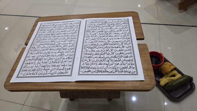 A Quran rests open on a wooden stand beside a small tray holding what appears to be dates and pickled vegetables. The caption playfully reframes the religious image—traditionally associated with spiritual blessings and divine gifts—as a humorous "airdrop" meme, suggesting the user has received unexpected rewards with "no capital" and quick, effortless returns.