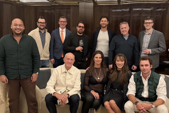 A group of eleven well-dressed individuals pose together in what appears to be a Manhattan social setting, with one man in a "Bitcoin" graphic t-shirt visible among the gathering, reflecting the post's celebration of Bitcoin in NYC.
