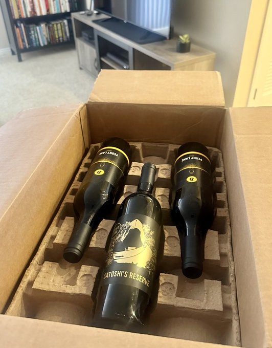 An open cardboard box reveals three black wine bottles with gold labels, one standing upright reading "SATOSHI'S RESERVE" and two lying down reading "RARE SATOSHI".
