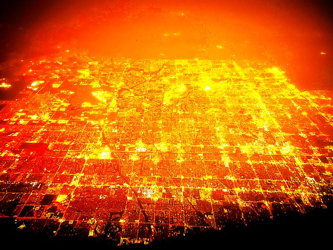 An aerial nighttime view of a sprawling city illuminated in intense red and orange light, creating a fiery, apocalyptic atmosphere.