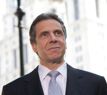 A man in a dark suit, white shirt, and light blue patterned tie stands outdoors with buildings blurred in the background.