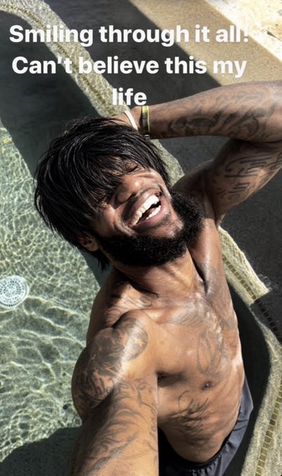 A tattooed man poses shirtless by a pool, with overlaid text reading 'Smiling through it all! Can't believe this my life'.