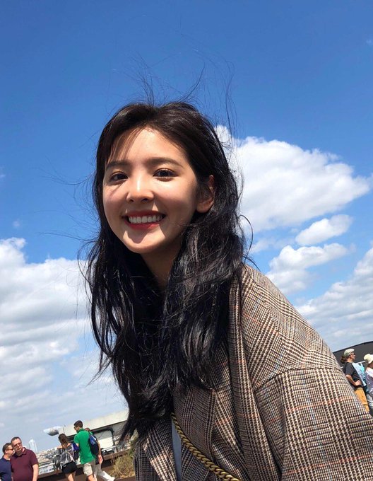 A young woman with long dark hair smiles for a selfie outdoors on a sunny day with a blue sky and clouds in the background.