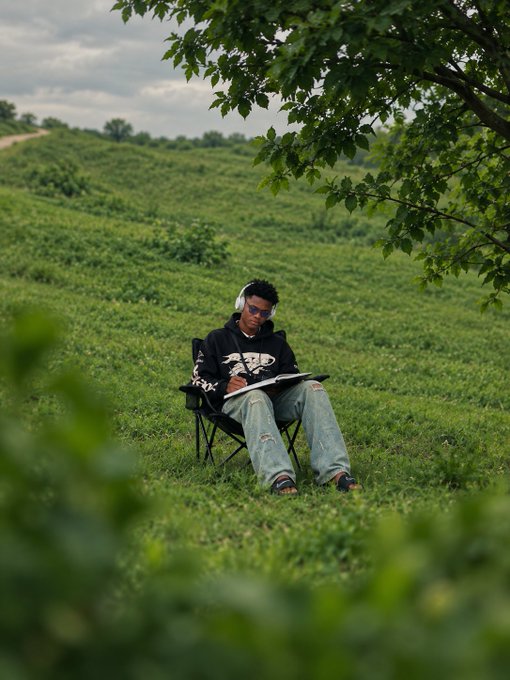 A person sits in a folding chair in a lush green field, writing in a notebook while wearing headphones and a black hoodie.