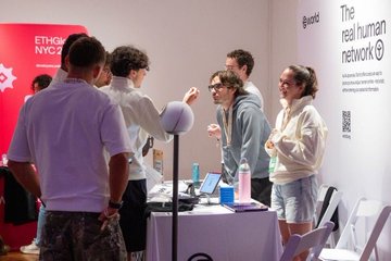 The image shows a group of people gathered around a table at an event. There are six individuals standing near the table, engaged in conversation with each other. The table has various items on it, including a laptop, a mouse, and several cups scattered across its surface. A sign is also visible in the background, which reads "The Real Human Network".
