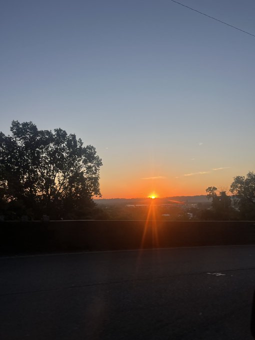 A golden sunset breaks over a treeline and distant hills, casting warm rays across a quiet roadway in silhouette. The image captures the imagery of "out of darkness into His marvellous light," with the rising sun symbolizing the radiant hope of 1 Peter 2:9 shared with the GM family.