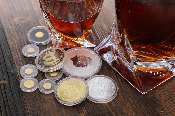 Photograph of two glass tumblers containing amber liquid (whiskey/bourbon) sitting on a wooden table alongside physical Bitcoin commemorative coins in gold and silver, plus a chocolate coin.