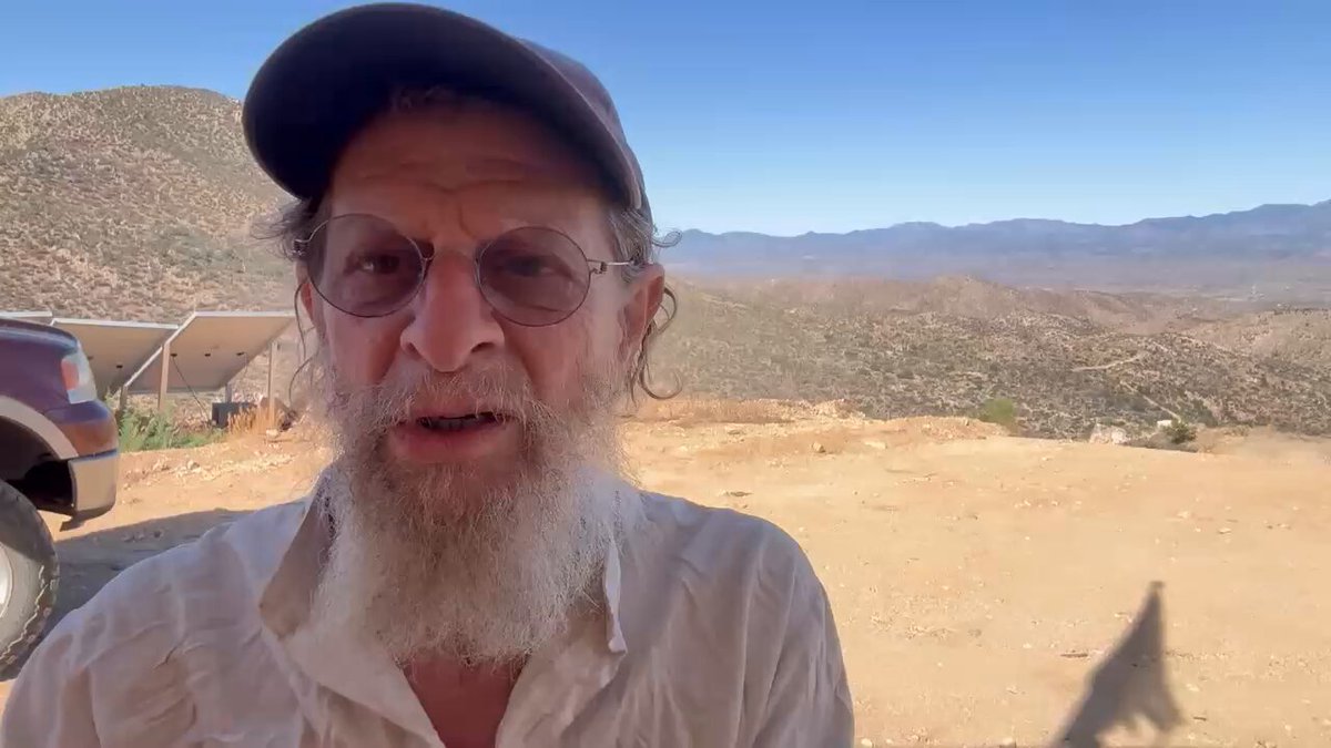 The image features a man wearing glasses and a hat standing in front of a desert landscape with mountains in the background, possibly at Fort Knox. The man appears to be talking or posing for a photo while looking directly into the camera. The caption provided seems unrelated to the main subject matter but includes some interesting phrases like "XRP, XLM, silver, gold, Uphold, Fort Knox will be audited. The Satanic Banking system is being destroyed. Buy Land. Leave the Matrix." This could possibly be a part of an advertisement or promotional material for a cryptocurrency exchange or related business.