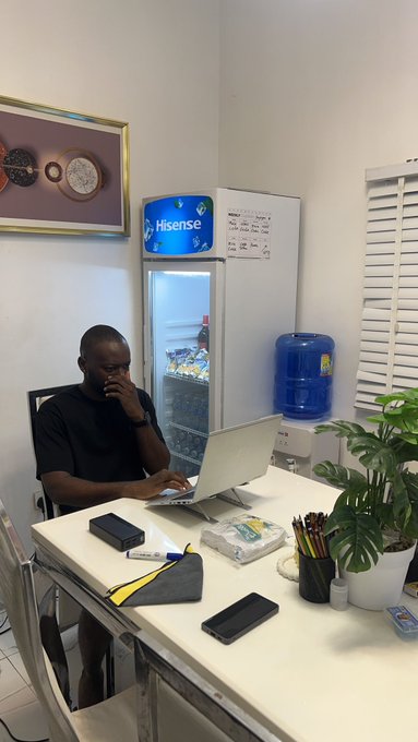 A man sits at a desk working on a silver laptop next to a Hisense refrigerator and a water dispenser in a small office space.