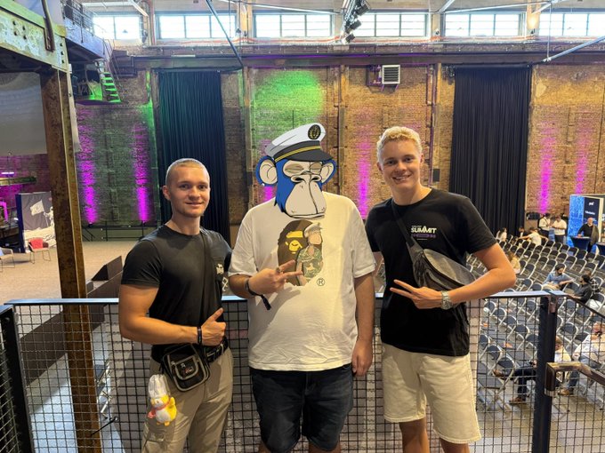 Two young men pose next to a person wearing a monkey mascot mask and a BAPE shirt at what appears to be an indoor event space with stage lighting.