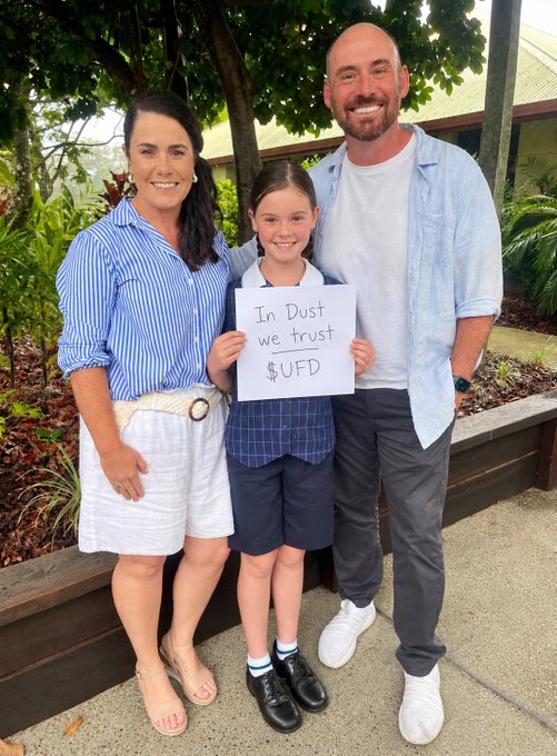 A family of three poses together outdoors, with the young girl in the middle holding a sign that reads 'In Dust we trust $UFD'.