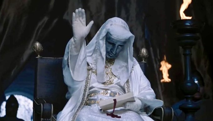 A hooded figure with blue-tinted skin sits on an ornate throne, raising one gloved hand while holding a scroll in the other, surrounded by flickering torches in a dim cavernous setting. The image's imposing, ceremonial atmosphere complements the post's reverent tone, visually representing a commanding "founder" figure addressing followers in solemn silence.