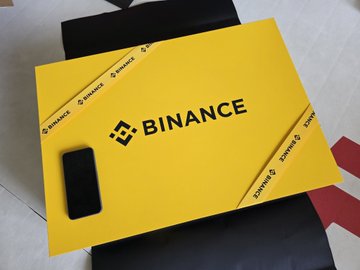 The image features a yellow box with a black border that says "BINANCE" written in white letters. The box has a cell phone placed inside of it, indicating that it contains electronic devices or peripherals. The box appears to be a gift from Binance, and it is being sold for 120 yuan on an online marketplace called Xianyu. This suggests that the image is related to a promotional post about Binance's peripherals, likely highlighting their luxury-grade quality and the platform's focus on users' satisfaction.