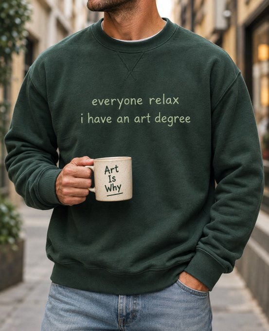 A man wearing a dark green sweatshirt reading "everyone relax i have an art degree" holds a mug that says "Art Is Why" while standing on a cobblestone street.