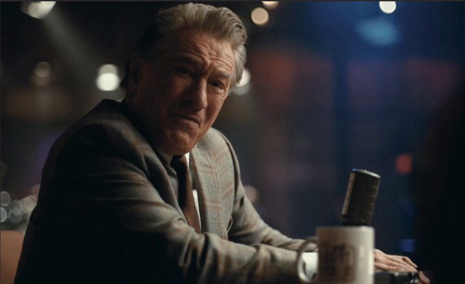 A still of an older man in a gray plaid suit sitting at a dimly lit bar or poker table with a bottle in front of him, set against a dark background with bokeh lights.