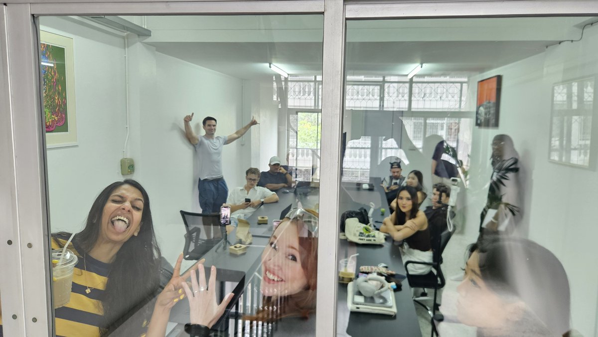 A group of people are seen through a glass window in what appears to be an office or meeting room, with one woman in the foreground laughing and sticking out her tongue.