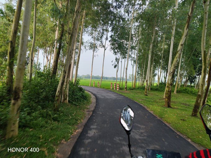 A first-person view from a motorcycle riding down a paved path lined with tall trees on both sides, with a digital speedometer visible showing '25' and 'HONOR 400' in the corner.