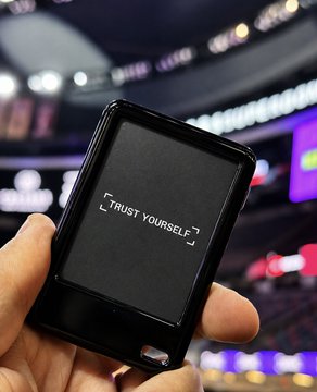 A hand holds up a Ledger Nano hardware wallet displaying "TRUST YOURSELF" on its screen, awarded as a MoonPay x Games League prize to the new owners. The blurred arena backdrop suggests the giveaway took place at a gaming or esports event.