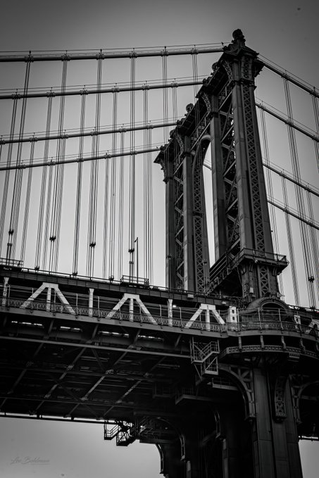 A black and white photograph of the Manhattan Bridge in New York, showcasing its suspension cables, stone tower, and steel architecture from a low angle.