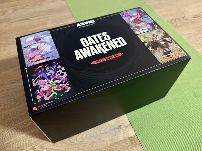 An Azuki TCG 'Gates Awakened - Relic Hunter' booster box rests on a green placemat over a wooden table, featuring anime-style character artwork on the lid.